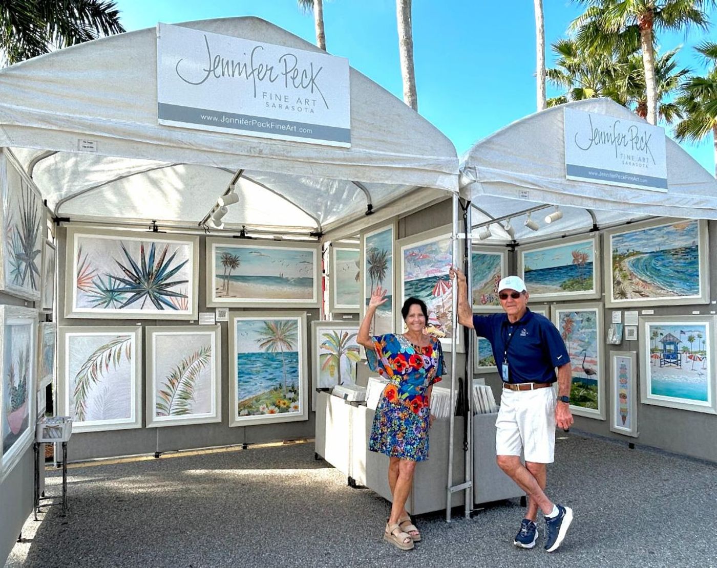 Jennifer waving beside Steve at their double booth — white tents with Jennifer Peck Fine Art banners and framed coastal collages