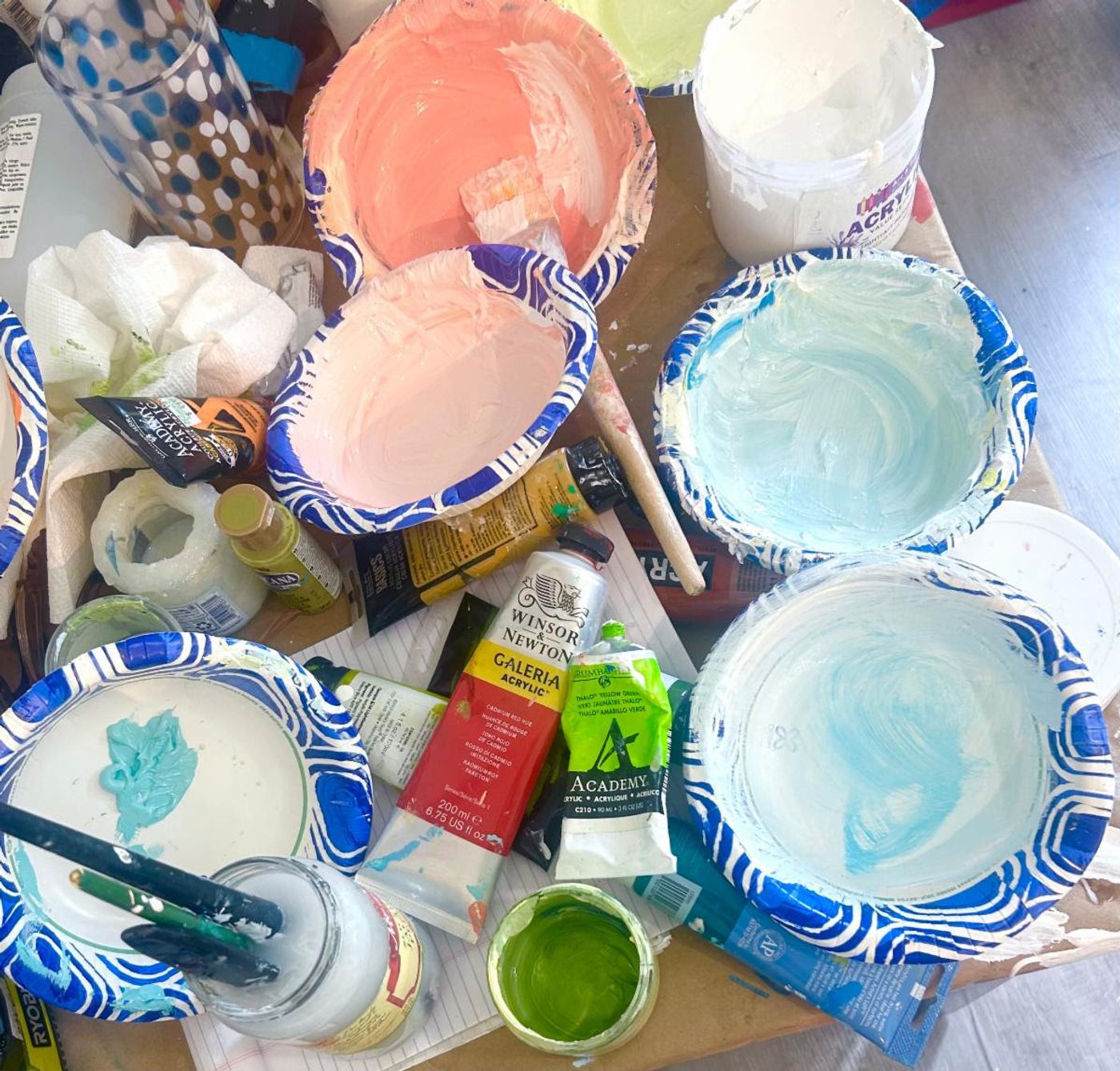 Paper bowls of coral, blush, and pale blue acrylic mixes among paint tubes on the studio table