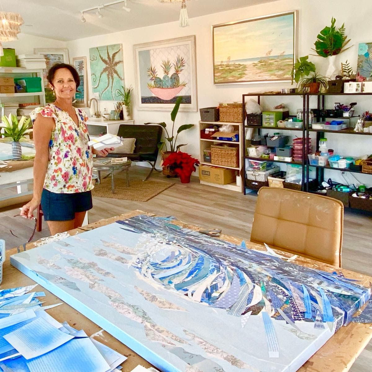 Jennifer in her bright studio beside a large wave collage in progress, blue paper strips layered across the canvas on her worktable