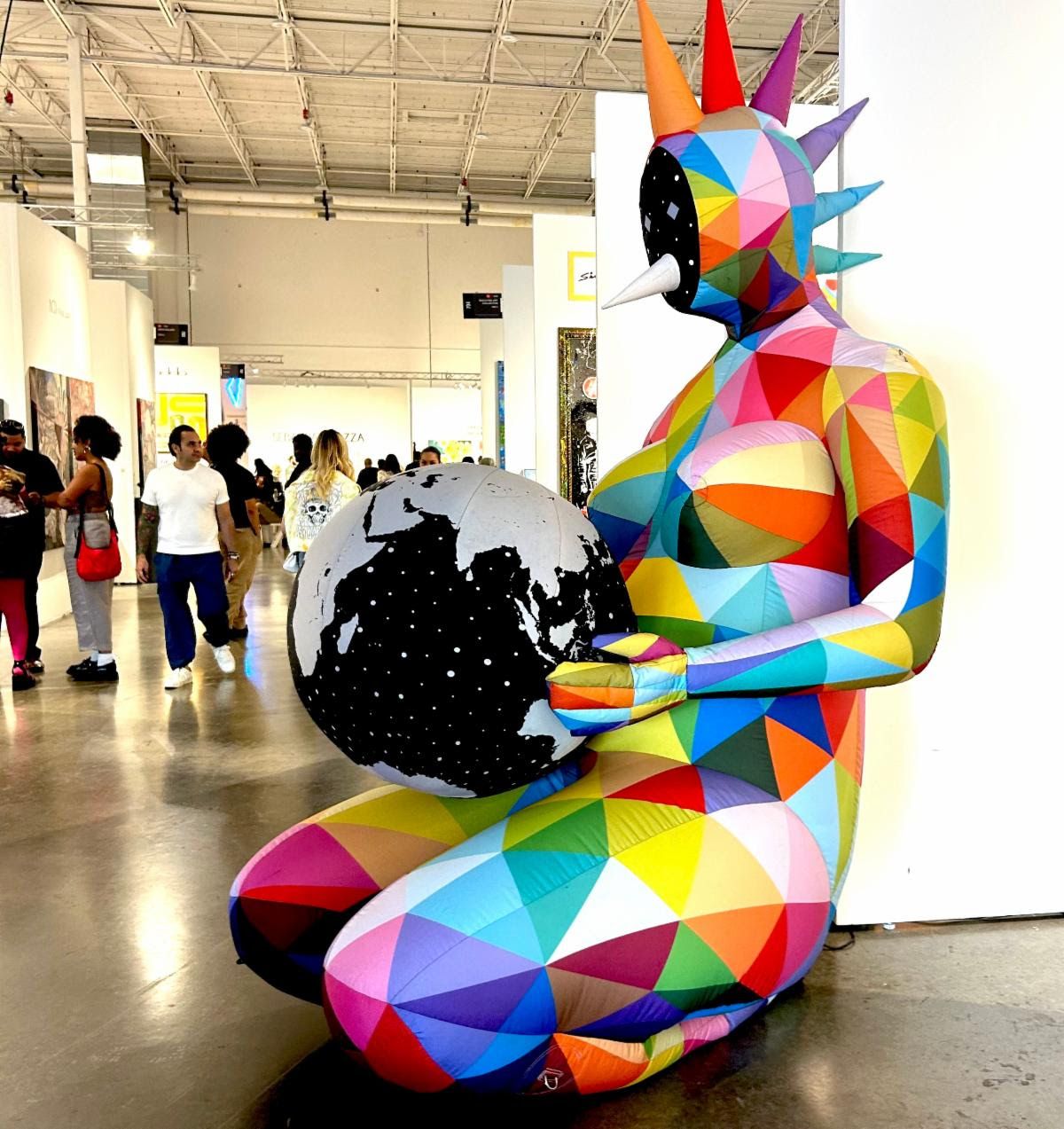 A rainbow-patchwork inflatable figure with a spiked crown kneels holding a black-and-white globe in a Miami fair aisle.