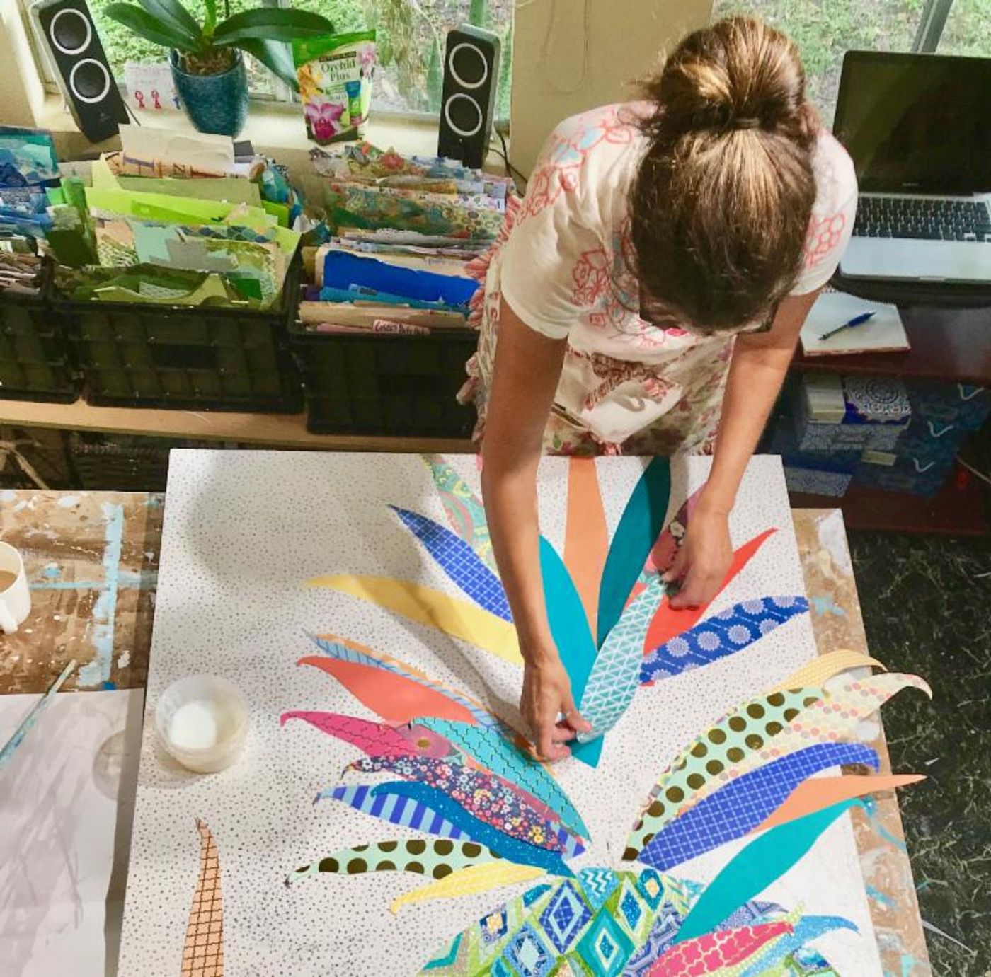 Jennifer leaning over a big square canvas, placing patterned cut-paper leaves for a colorful pineapple collage