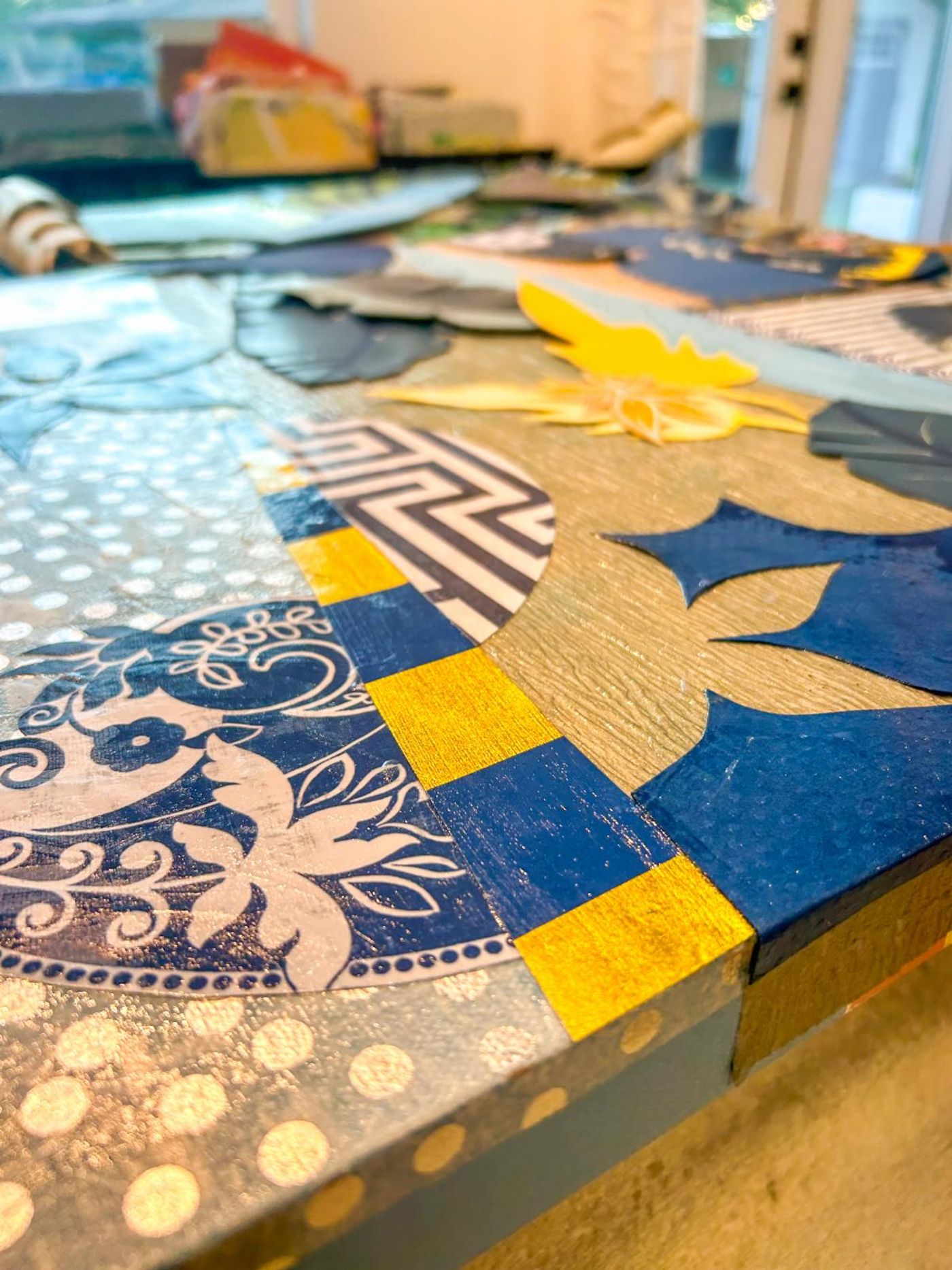 Angled close-up of a collage panel edge: blue-and-white floral medallion, gold-and-navy checked band and a navy leaf