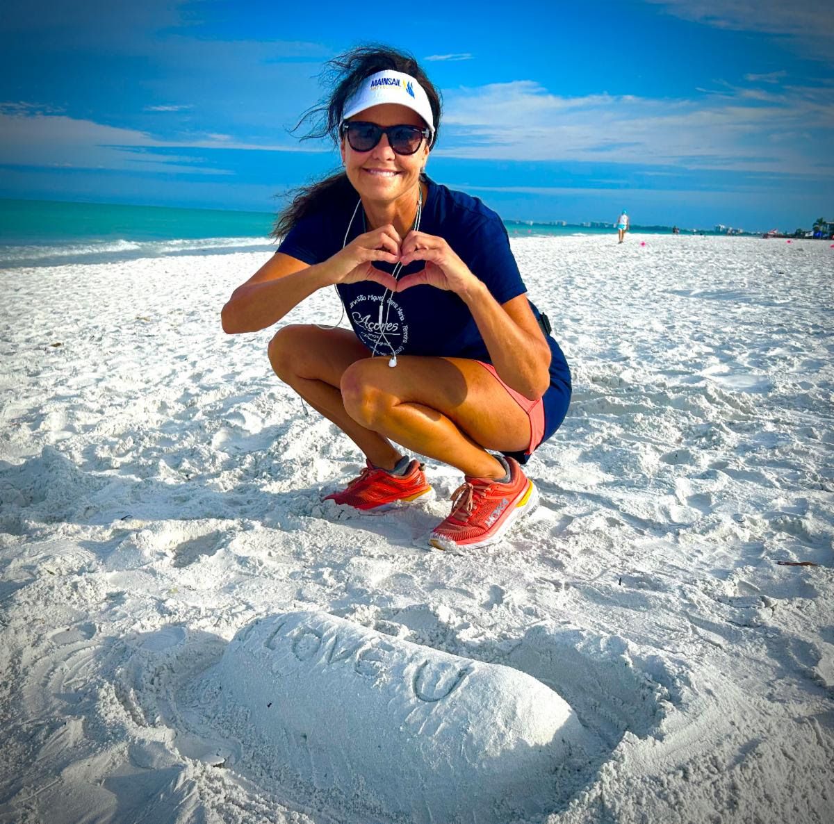 Jennifer makes a heart with her hands on bright white sand, 'Love U' carved into the beach in front of her