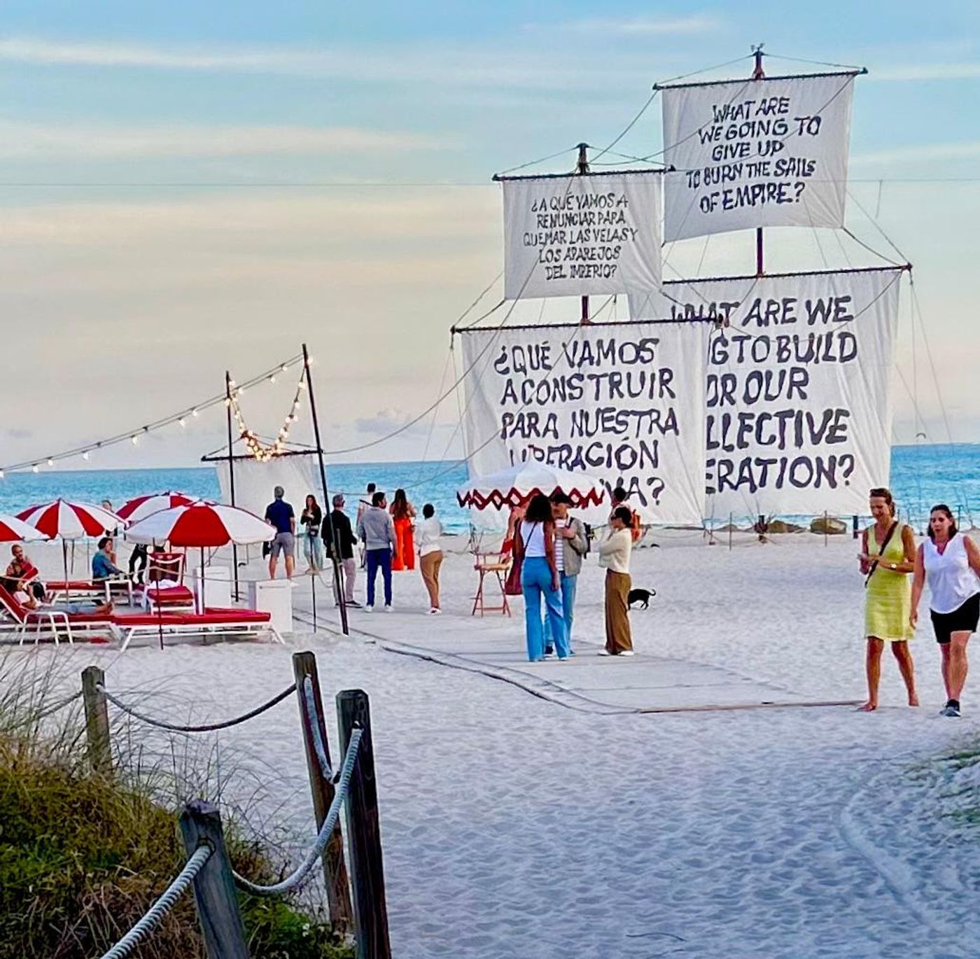 Ship sails hand-painted with questions in English and Spanish rise over Miami Beach, red-striped umbrellas below.