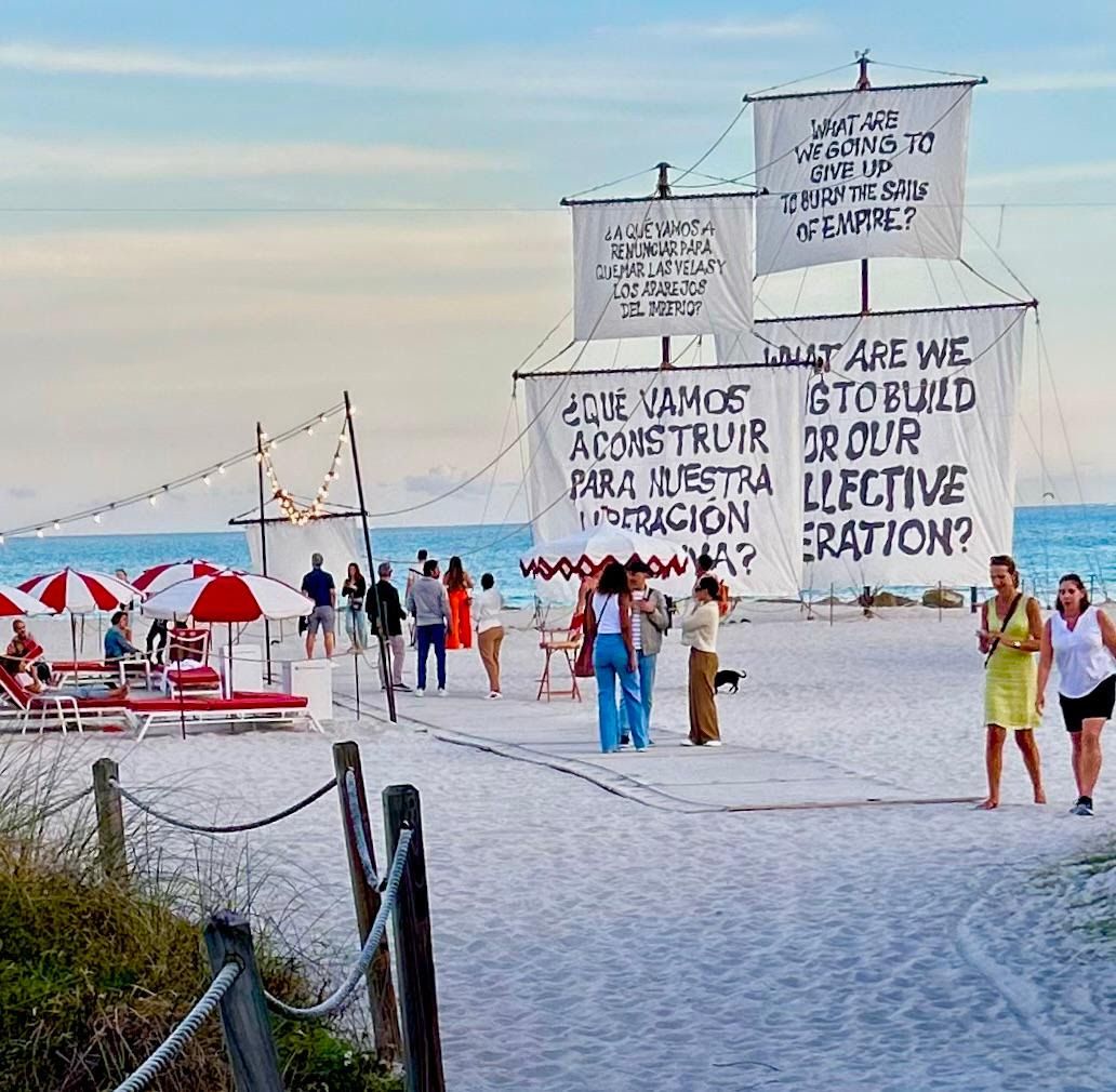 Ship sails hand-painted with questions in English and Spanish rise over Miami Beach, red-striped umbrellas below.