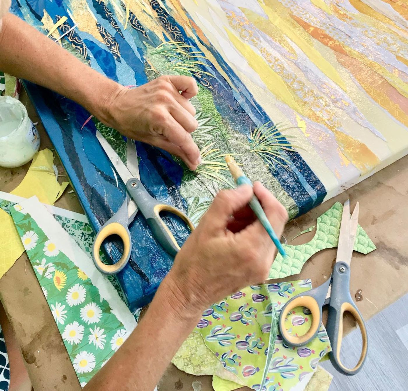Close-up of Jennifer's hands with a glue brush over the collage's deep blue water, scissors and patterned green papers nearby.