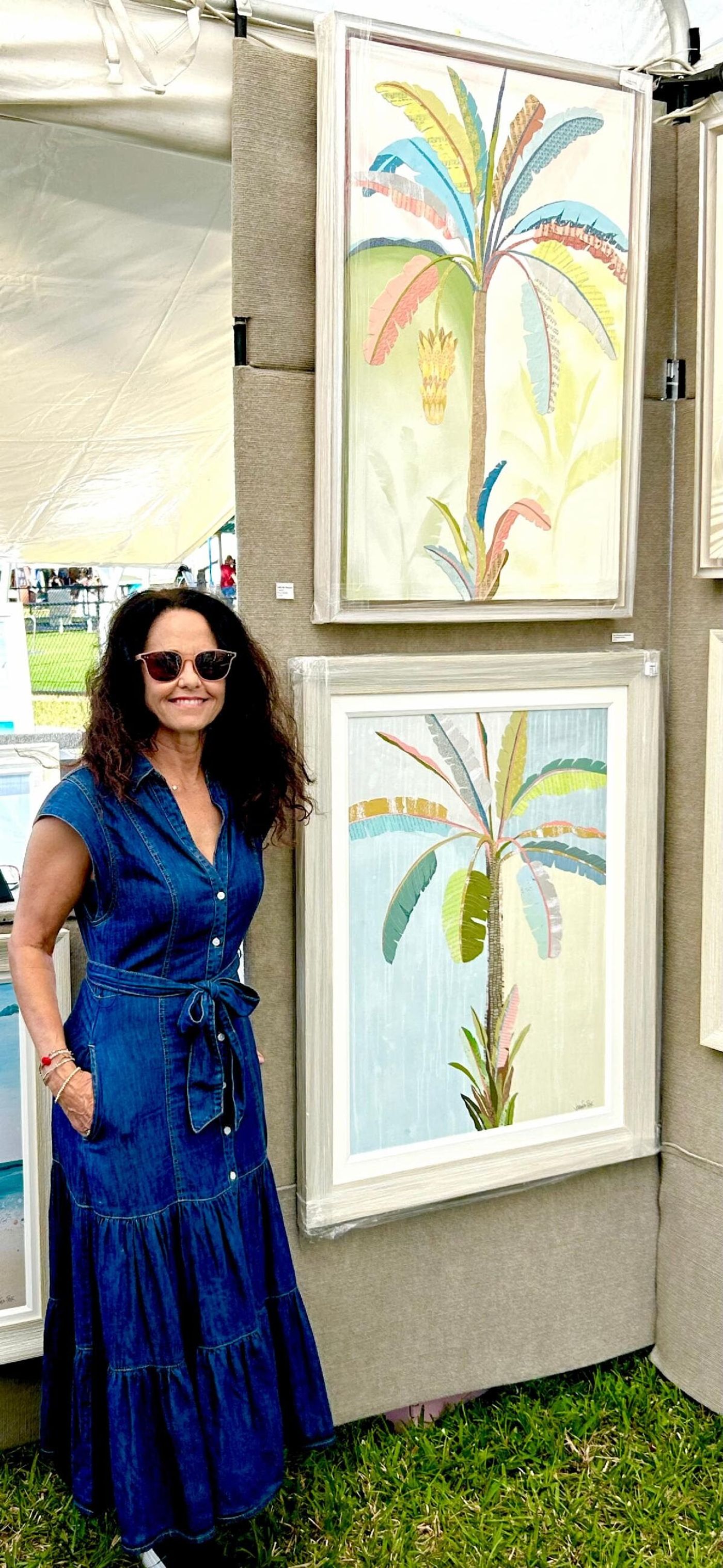 Jennifer in a long denim dress at her booth beside two framed banana palm collages, one with a hanging banana bunch