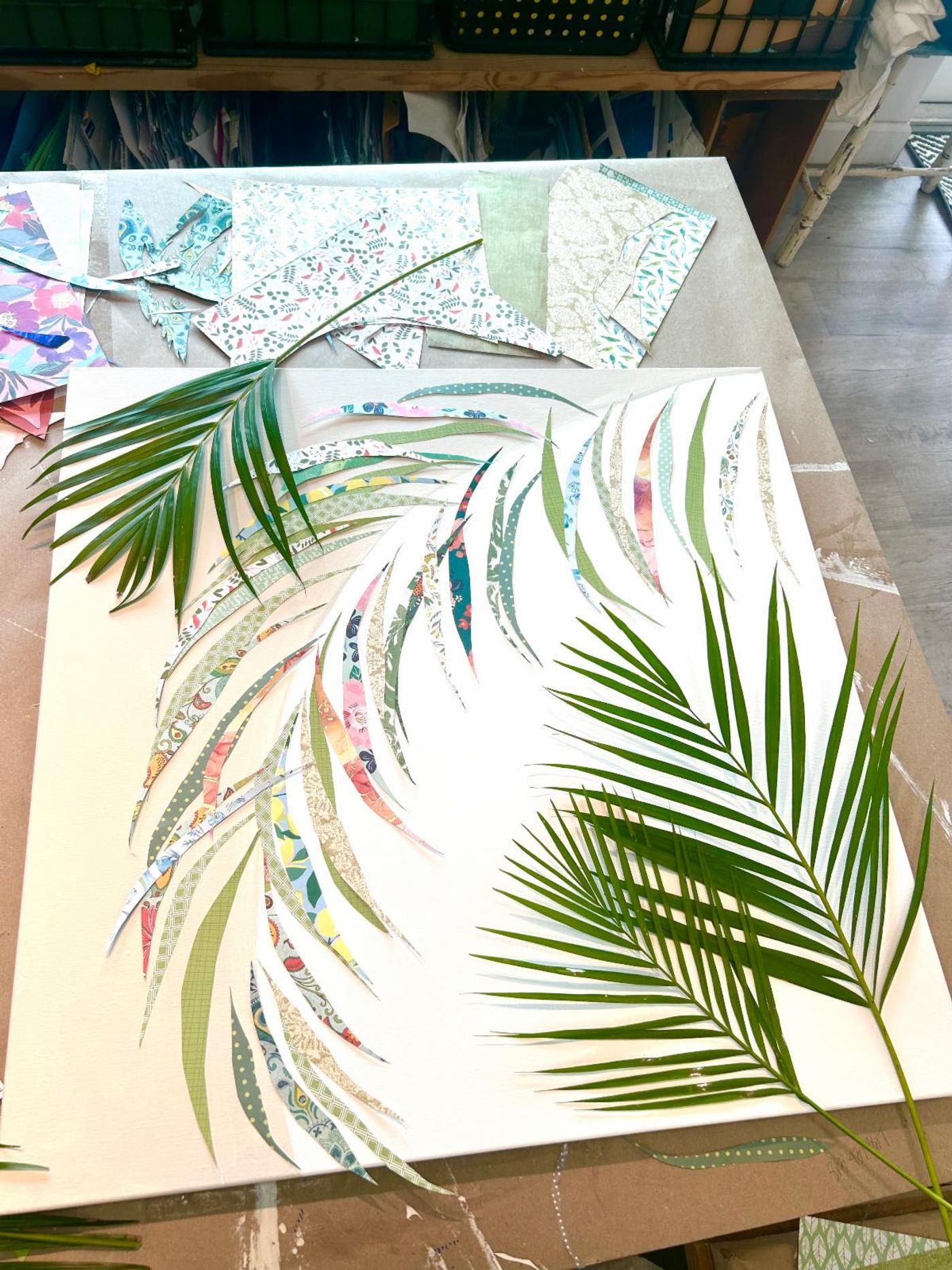 Two fresh palm fronds rest on a canvas where a frond of patterned-paper leaflets takes shape, paper scraps across the table.