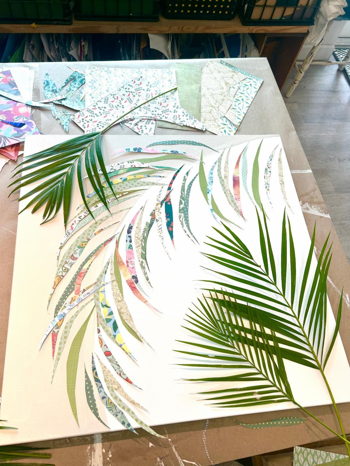 Two fresh palm fronds rest on a canvas where a frond of patterned-paper leaflets takes shape, paper scraps across the table.