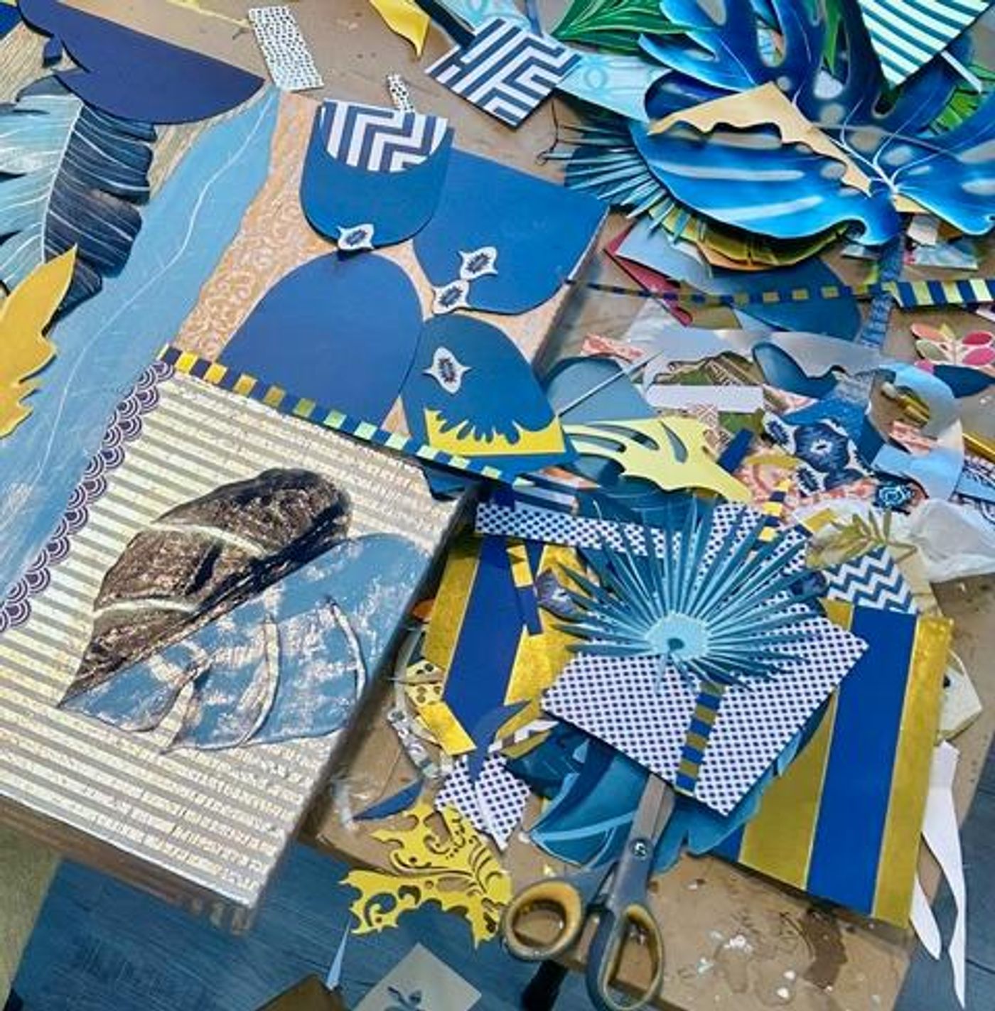 Studio worktable covered in blue and gold paper scraps, cut fans, leaves and chevrons, with scissors and panels in progress