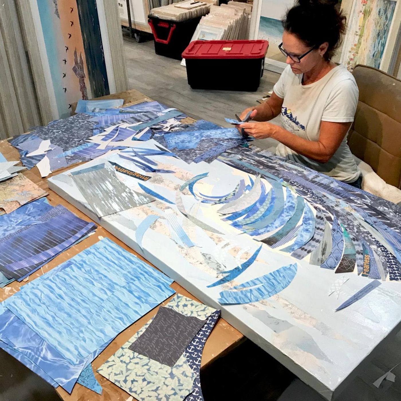 Jennifer at her studio table cutting blue papers as the Big Kahuna wave takes shape on the canvas beside her