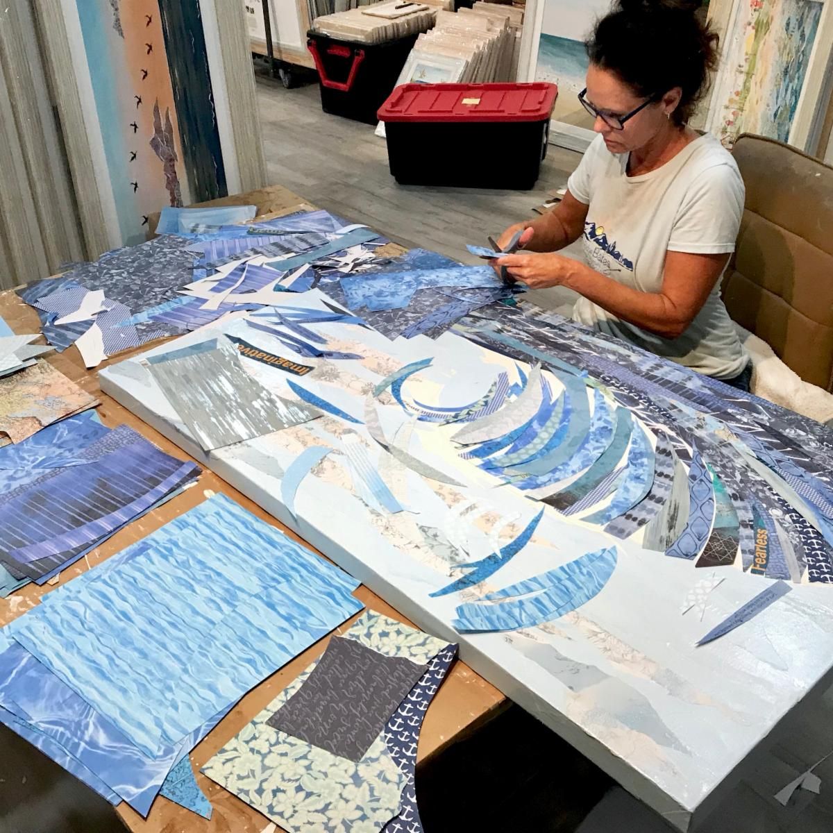 Jennifer at her studio table cutting blue papers as the Big Kahuna wave takes shape on the canvas beside her