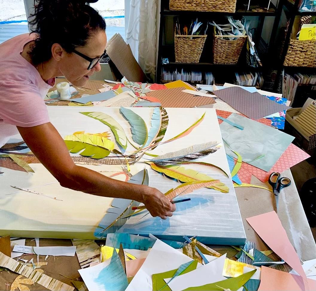 Jennifer places hand-cut paper leaves on a large banana palm canvas at her studio table, baskets of collage papers behind her