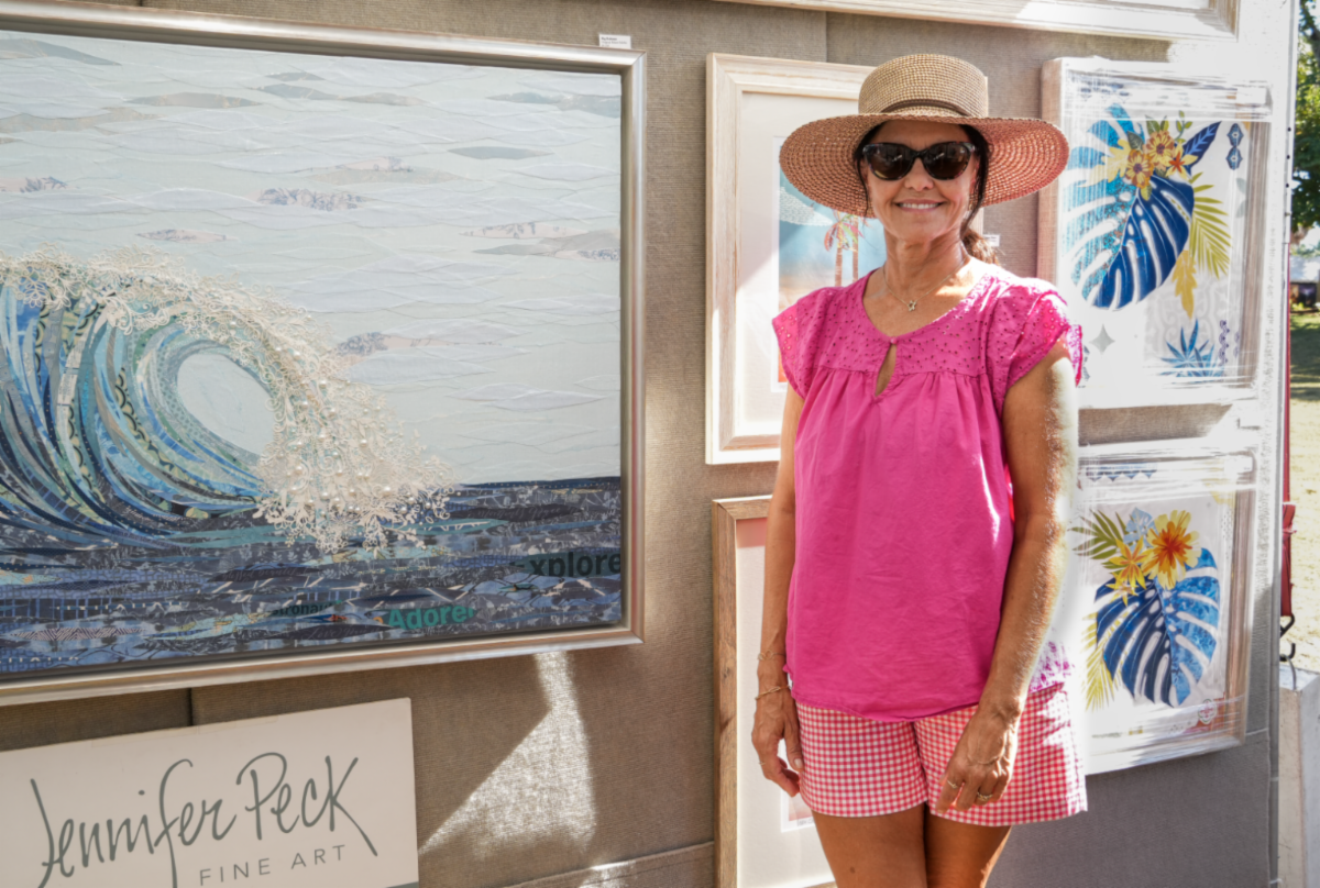 Jennifer in a straw hat and pink top at her booth beside a large cresting-wave paper collage, leaf collages hanging behind her
