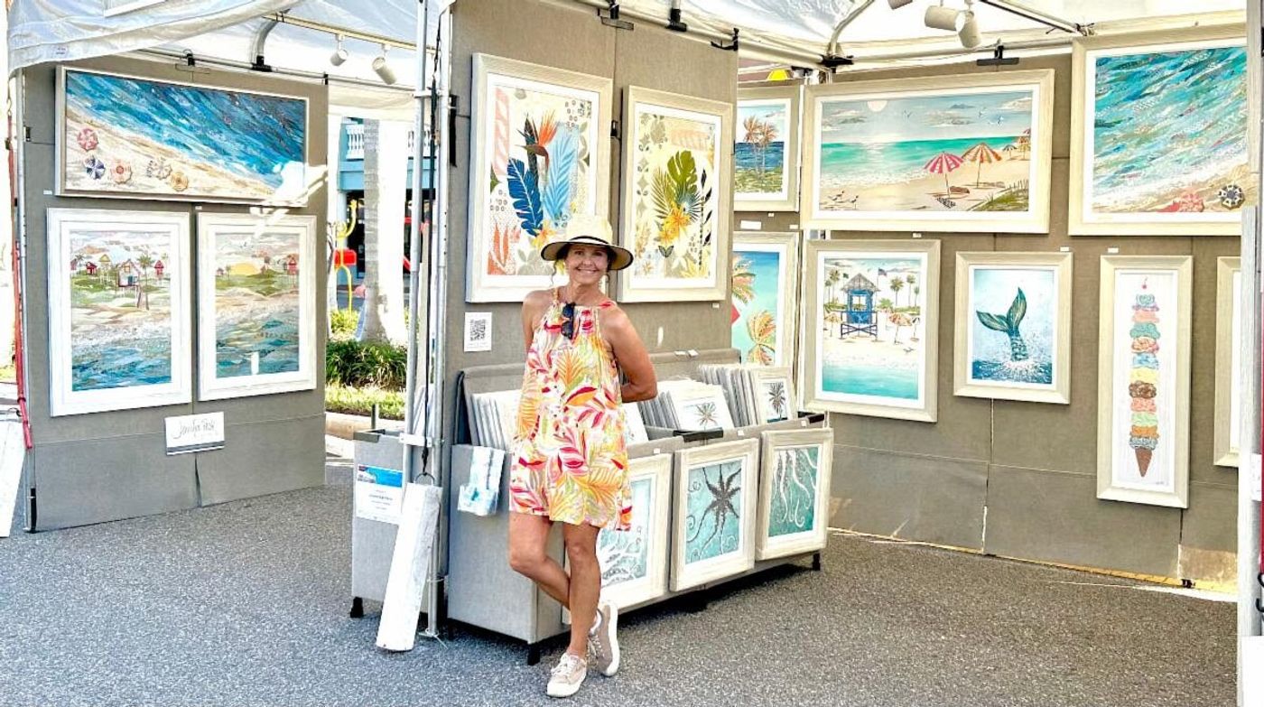 Jennifer in a sun hat at her festival booth, walls hung with framed coastal collages — beach scenes, a mermaid tail, an ice cream stack