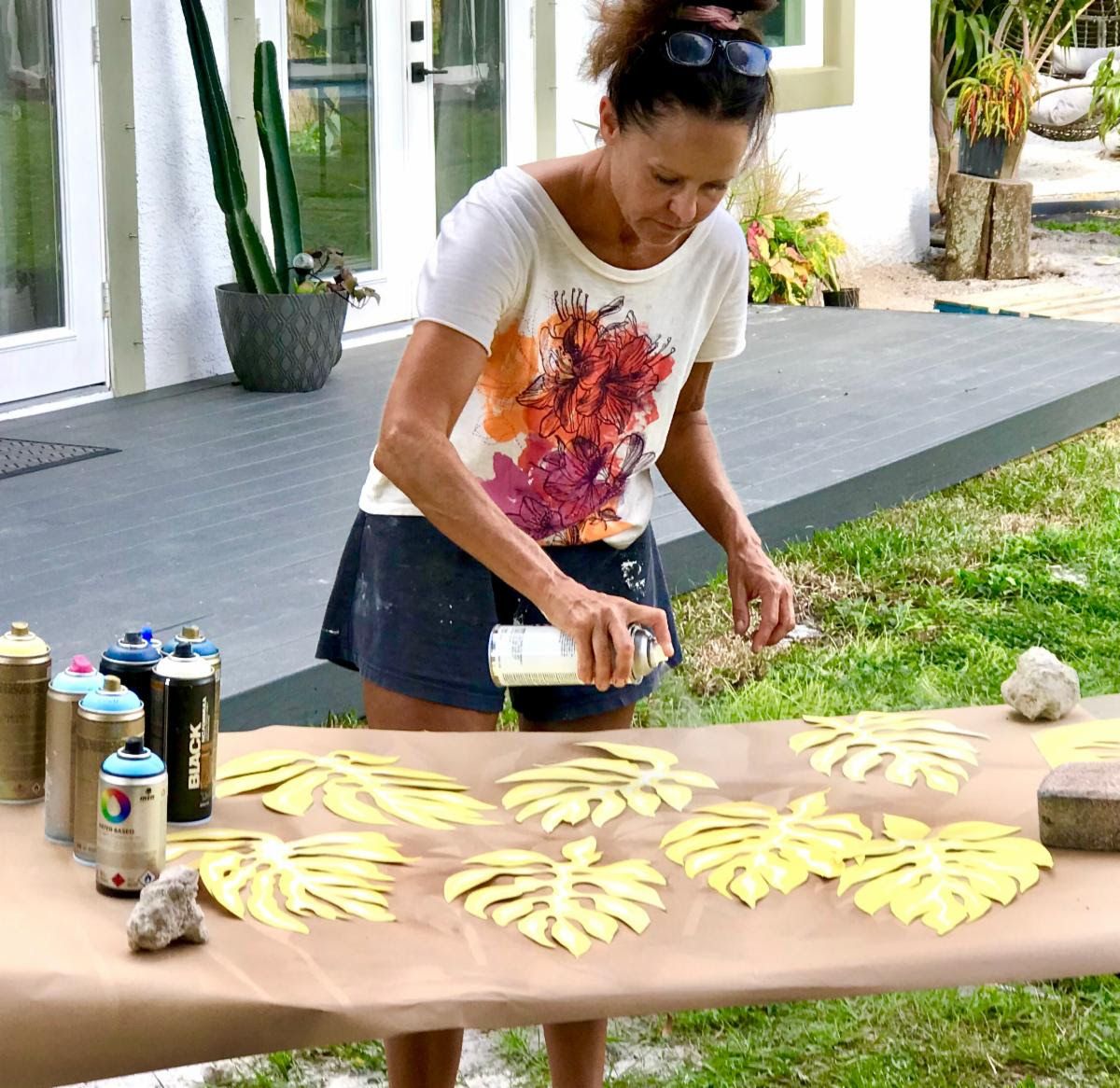 Jennifer spray-paints hand-cut yellow monstera leaves laid out on kraft paper outside the studio