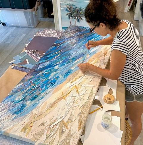 Jennifer leans over the long beach collage on her work table, gluing blue paper waves beside the finished sandy shore.