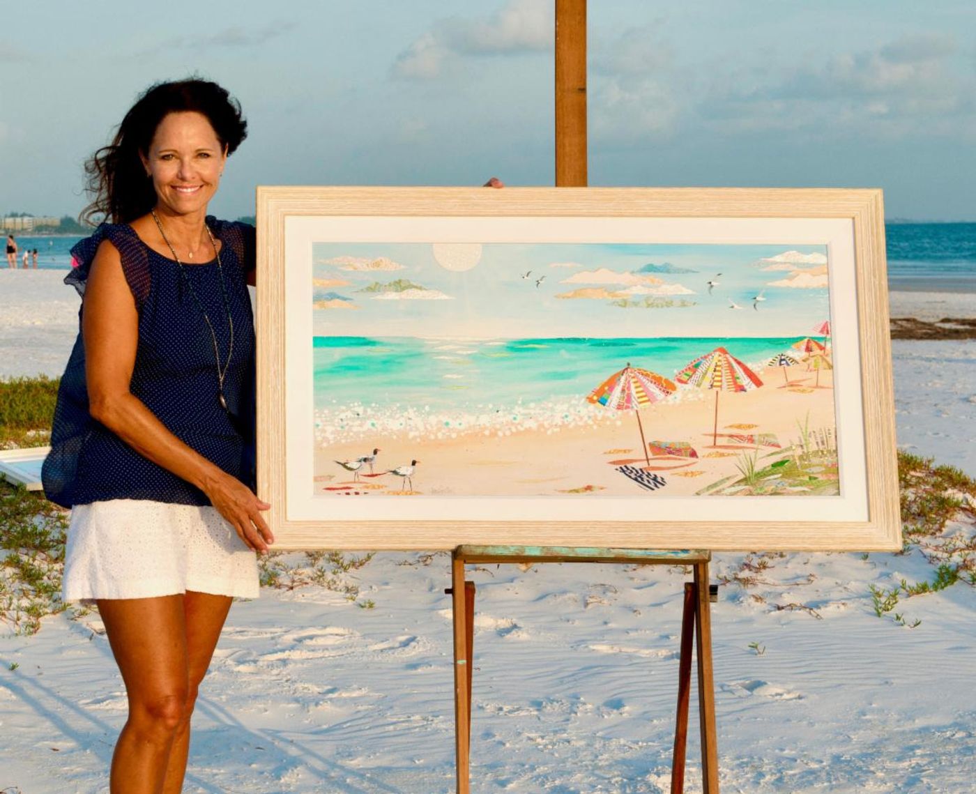 Jennifer on white sand beside a large framed beach collage on an easel — turquoise surf, striped umbrellas and gulls