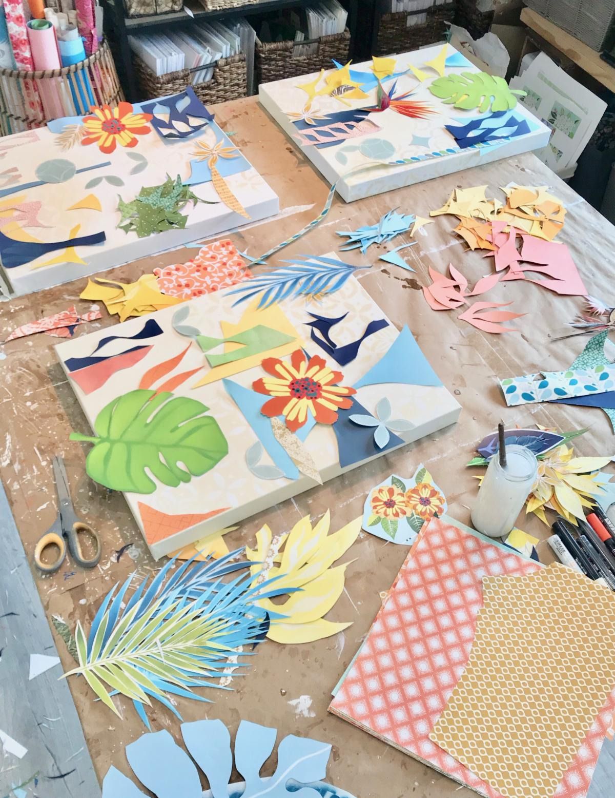 Three collages in progress on the studio table, loose cut-paper leaves and flowers arranged over warm cream backgrounds