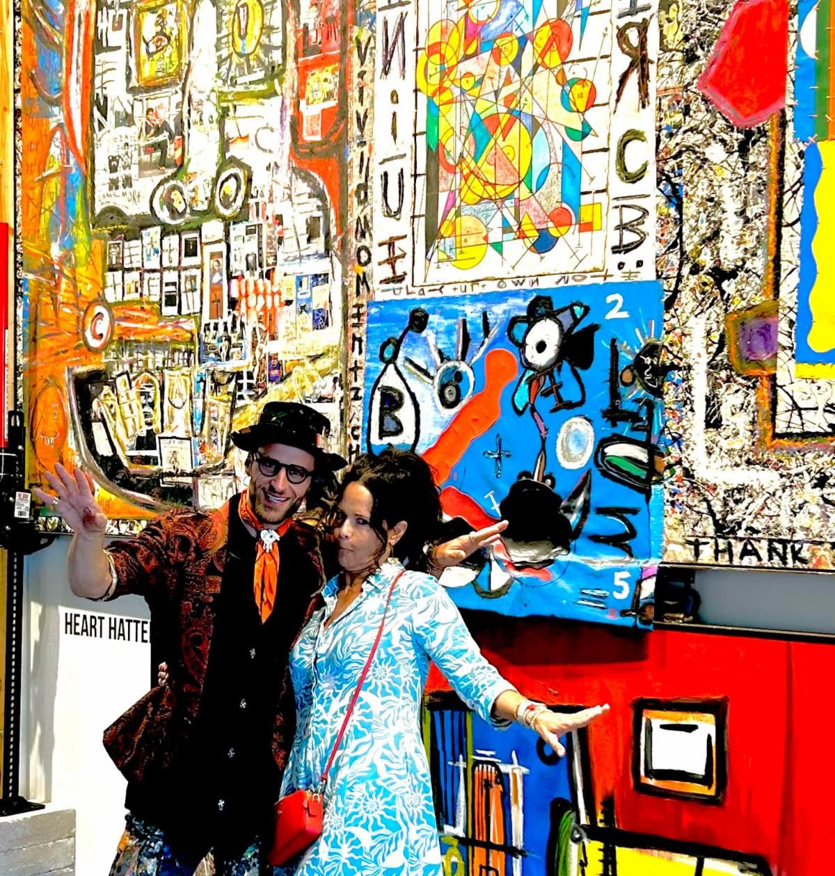 Jennifer and an artist in a black hat spread their arms wide before densely collaged paintings at the Heart Hatter booth.