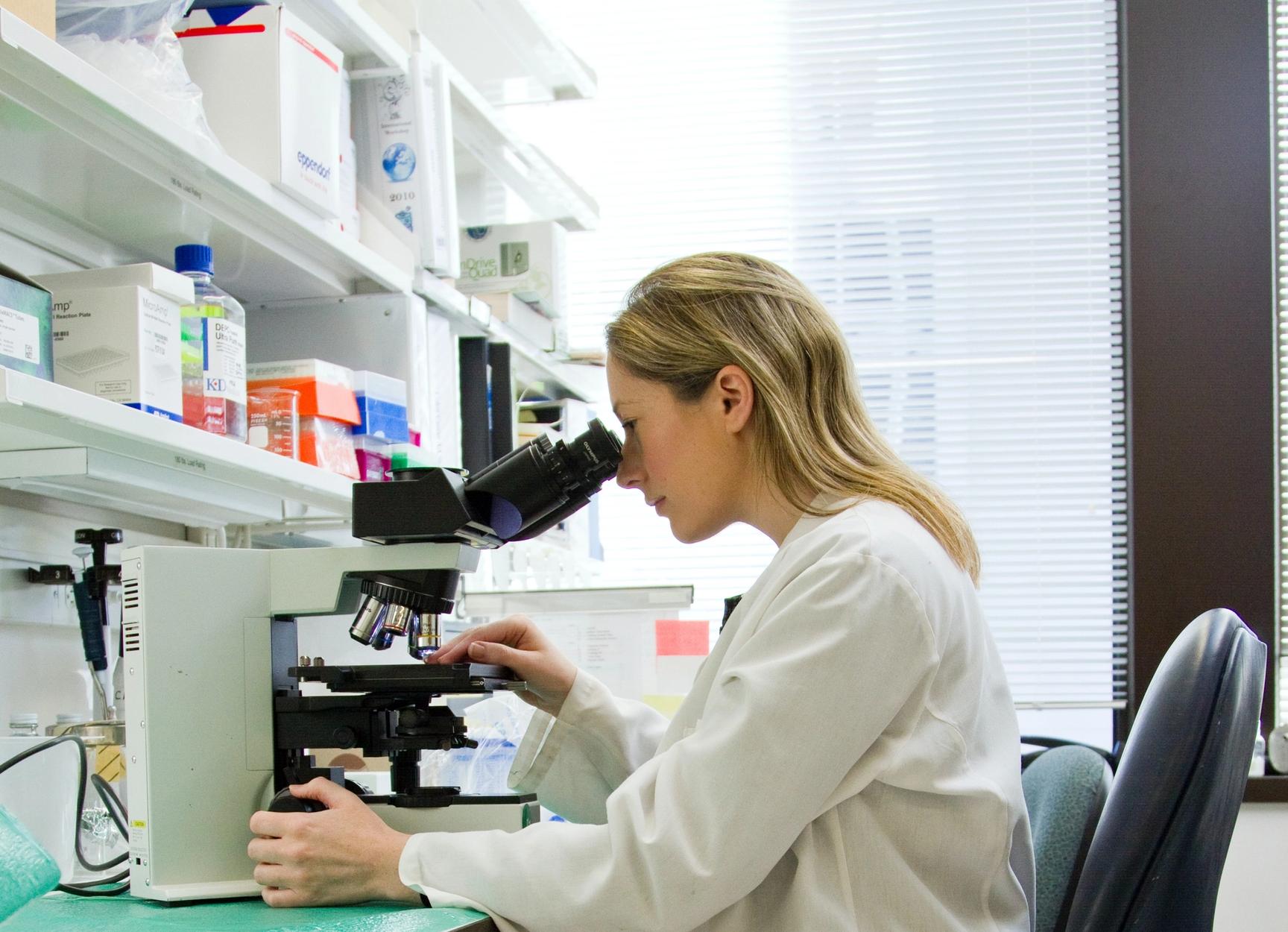 En kvinne i labfrakk titter inn i et microskop. Hun sitter på en kontorstol på et kontor med legemidler i hyllene.