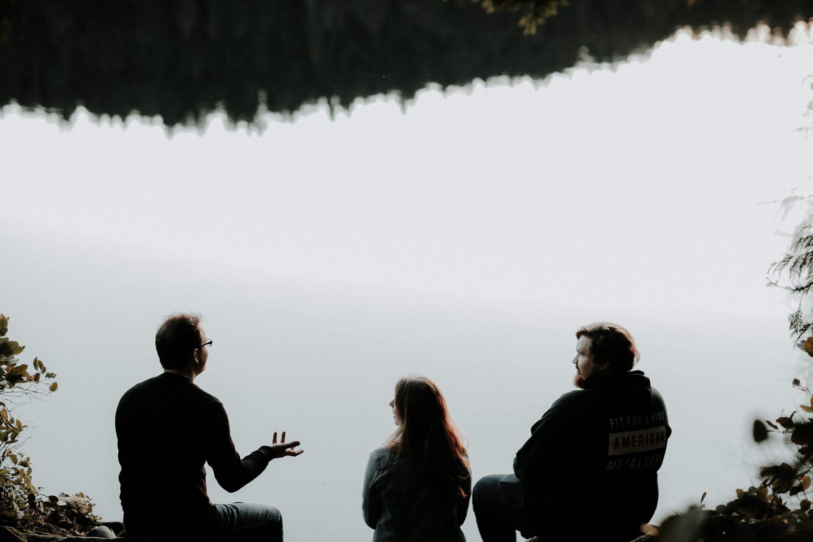 Tre personer snakker sammen. Vi ser dem bakfra og i bakgrunnen ser vi et vann som reflekterer det som virker å være en skog.