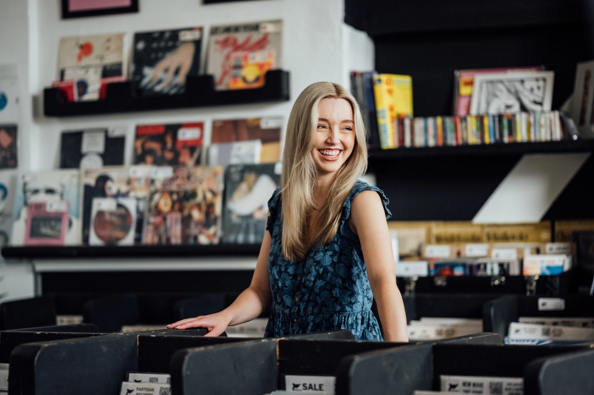A smiling woman in a blue dress stands among record bins and shelves of albums in a music shop.