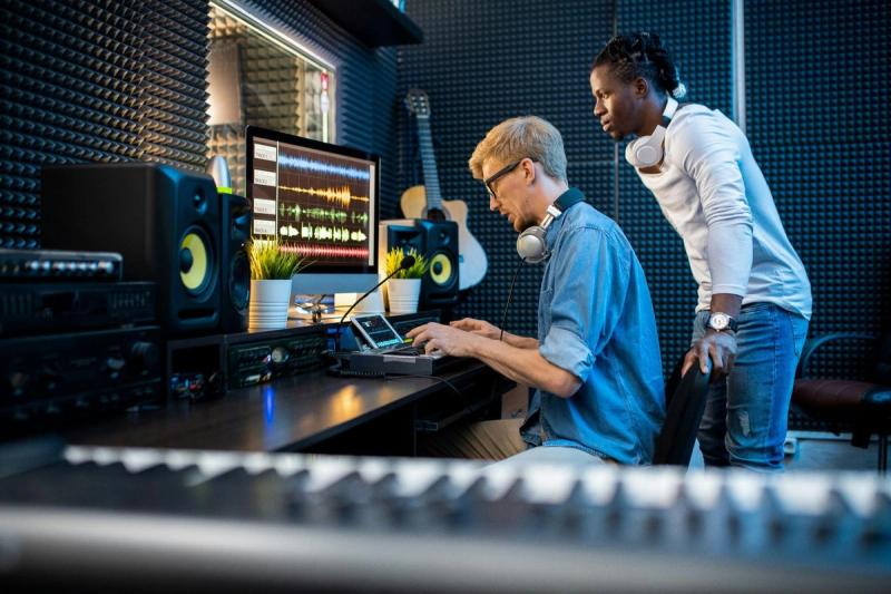 A mentor and student wear headphones while producing a song at a computer in a recording studio.