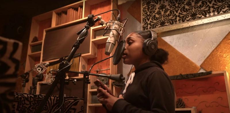 A young vocalist wearing headphones records vocals into a large studio condenser microphone.
