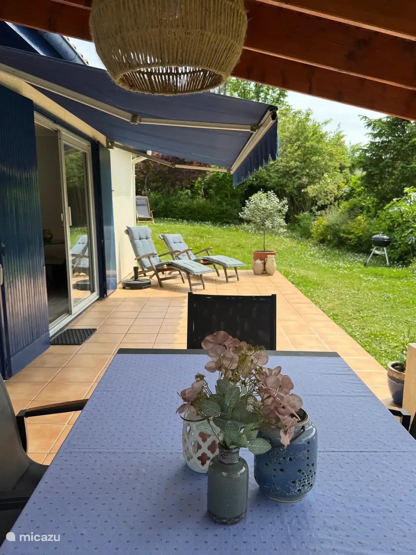 Het terras onder de blauwe zonwering, met uitzicht op de tuin en de bomen