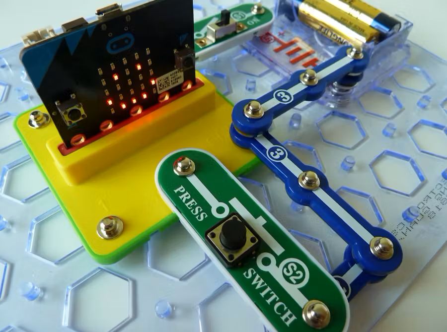 A micro:bit is docked to a Snap:bit board, attached to large plastic electronic components which work as wires, and a switch to make a smiley face appear on the micro:bit