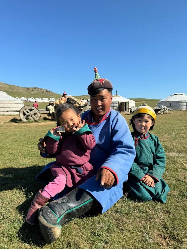 Smiling father and two daughters sitting on grass in Mongolia. One daughter is holding a micro:bit to the camera.
