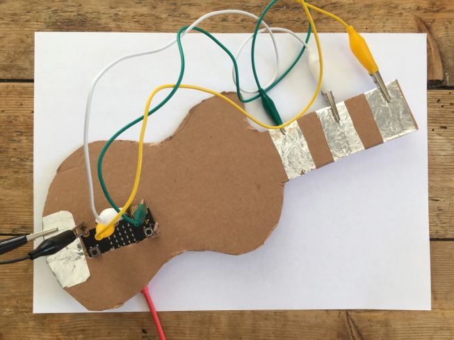 foto mostrando contactos de papel de aluminio en una guitarra de cartón conectadas a los pines 0, 1, 2 y GND del micro:bit 