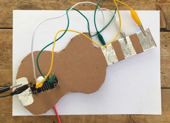 un micro:bit attaché à un morceau de carton en forme de guitare avec des pinces crocodile connectées à des sections couvertes d'aluminium sur le carton et les broches du micro:bit