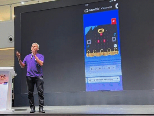 Mohammed Azrul Che Mustapha, holding a microphone and talking on stage with a micro:bit shown on a screen behind.