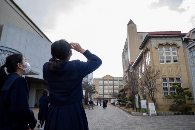 2 girls in school uniform are in a school playground looking upwards at the roof of the school