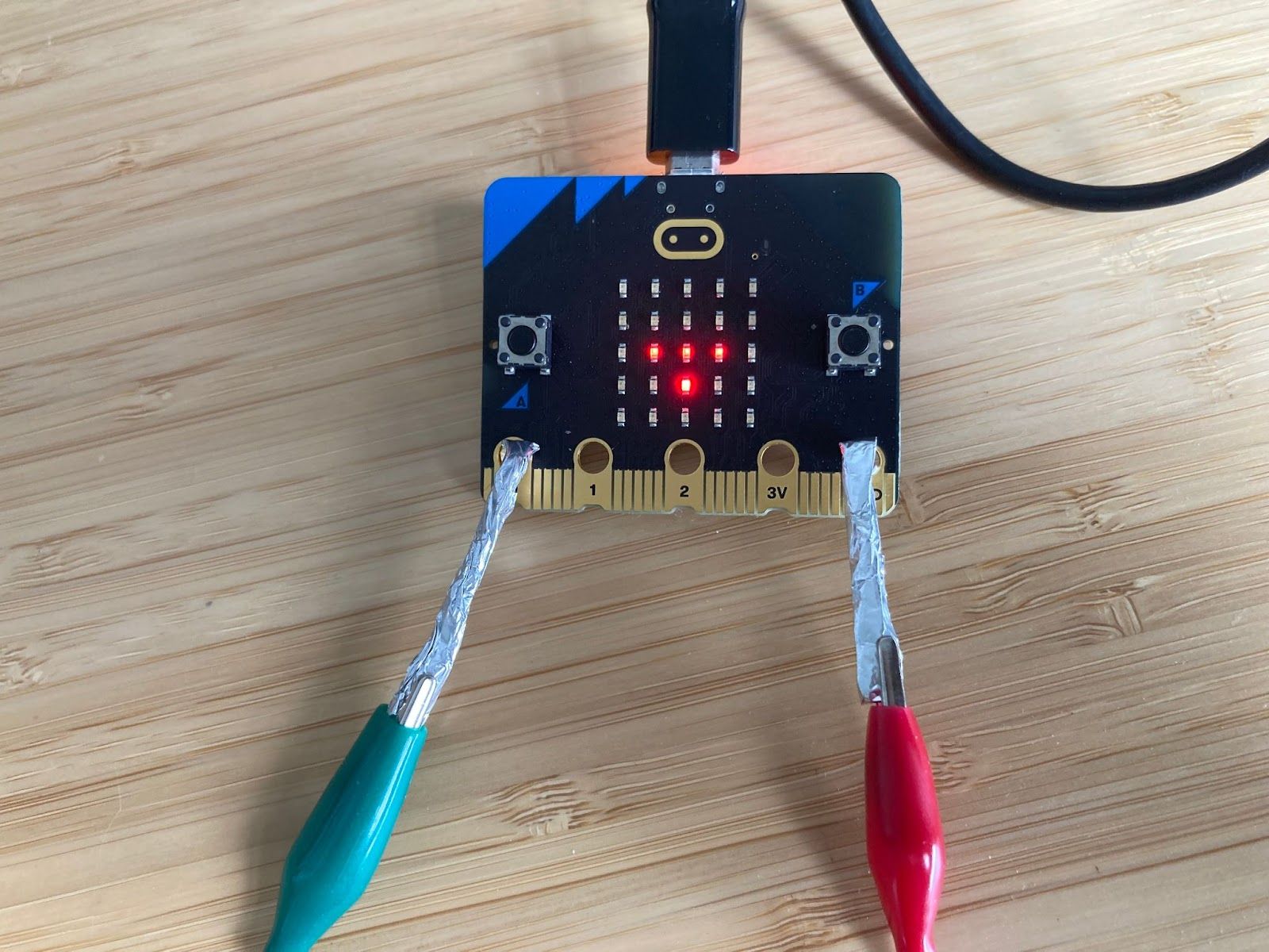 A micro:bit on a desk with green and red crocodile clips attached to pin 0 and GND using hooks made of aluminium foil.