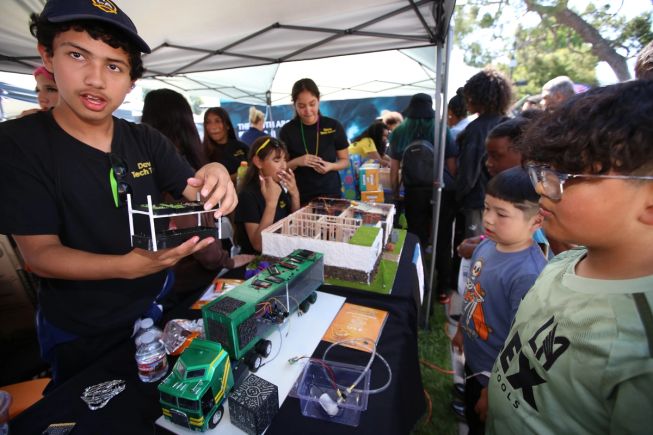Students demonstrating a technology project under a canopy tent at a STEAM event, with children and families gathered around to observe.