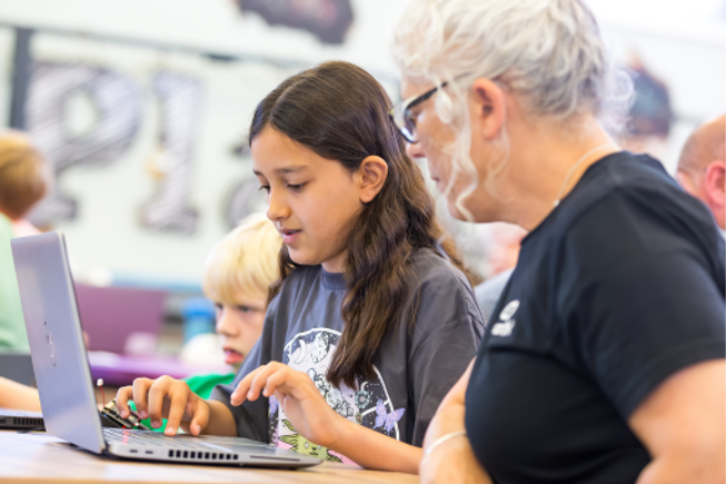 A young person works on a laptop alongside an adult, with other students in the background.