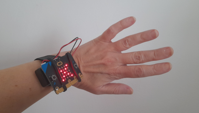 Photo of micro:bit with cross icon on its display, attached to someone's wrist.