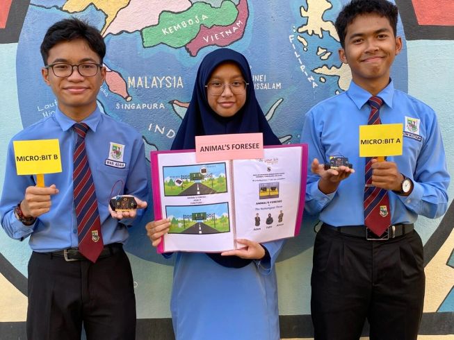 Image showing three children who took part in a do your :bit challenge, holding up their project idea and 2 micro:bits and standing in front of a world map.
