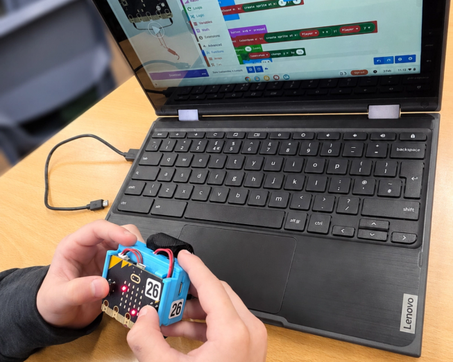 A pupil holds a micro:bit in a blue case beside a laptop displaying a MakeCode project.