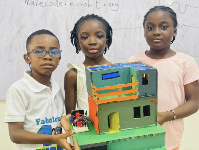 Three young people from Ghana stand behind a model house they created for their smart home security and safety project. The model includes a micro:bit sensor system designed to provide early warnings of fire, gas leaks and intruders.