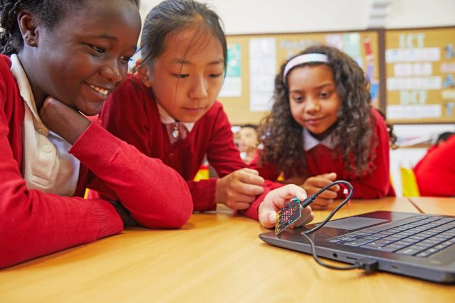 Three girls, primary school pupils, looking at a micro:bit that is attached to a laptop with a USB lead.