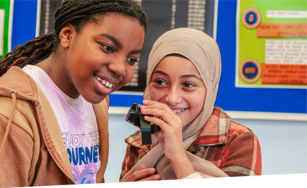 two smiling girls in a classroom, one holding a micro:bit device