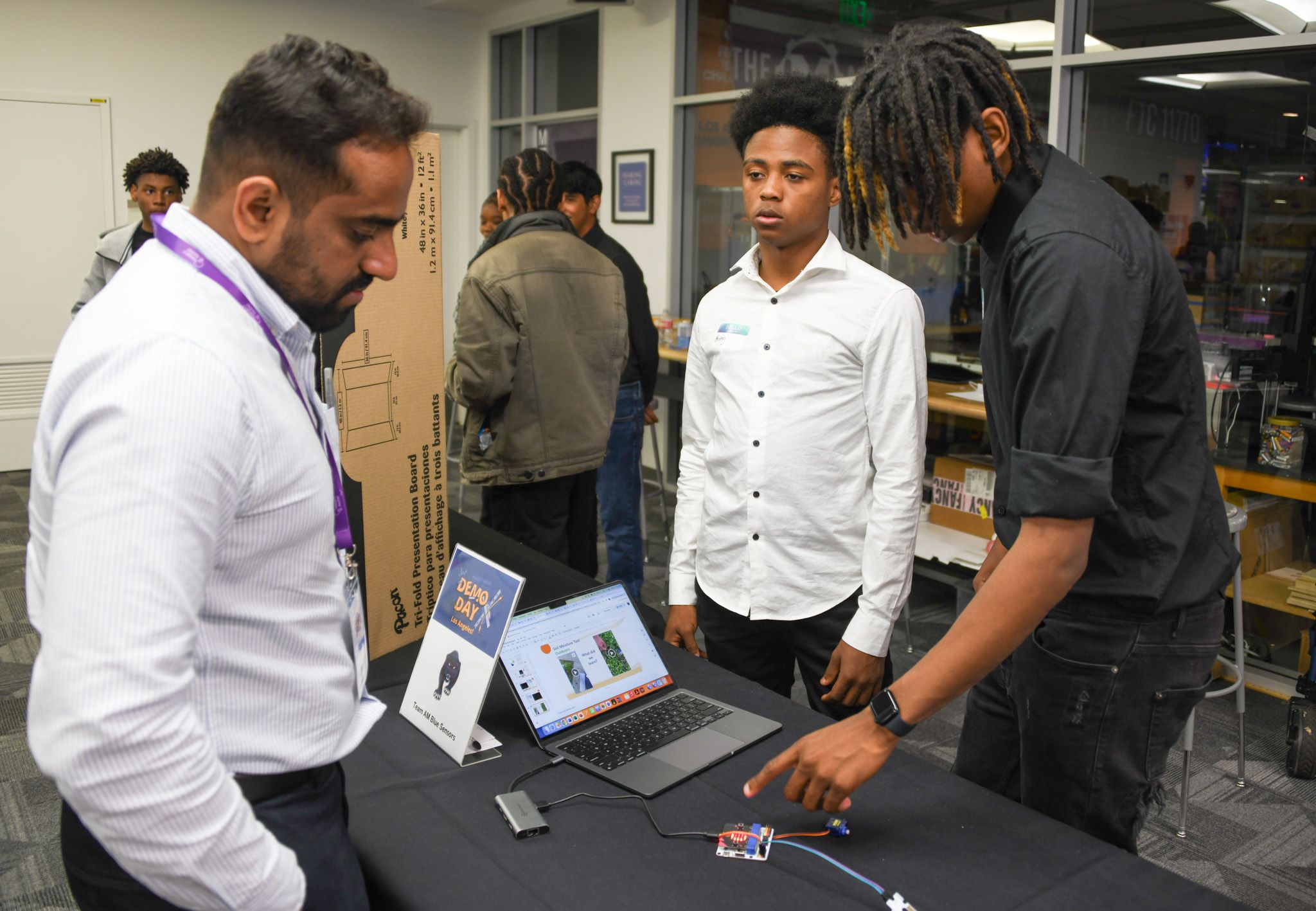Image shows students displaying their project and demonstrating it to a teacher.