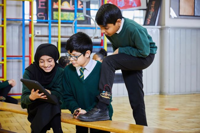 3 primary pupils use micro:bits in the school hall, including one micro:bit attached to a pupil's leg