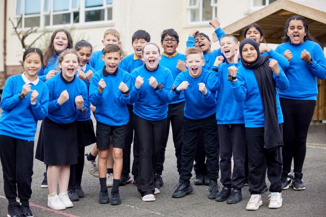 A group of students in royal blue jumpers and wearing micro:bits on their wrists cheer in their school playground