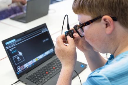A student using a micro:bit that has braille capital A and B labels above its buttons, connected via USB cable to a laptop.