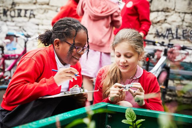 Two primary age girls are crouched down next to a planter in the playground looking at a micro:bit. One pupil is holding a clipboard and pencil.